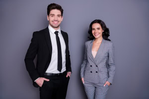 Portrait of beautiful man and woman with wavy curly brunette hair wearing black tuxedo jacket blazer isolated over grey background