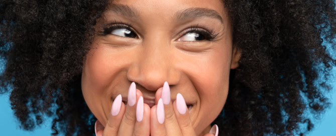 Close up of happy biracial girl Laughing dark skinned millennial woman looking away Cute positive mixed race female smiling, covering her mouth and lips with hands, blue background