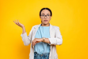 Confused puzzled hispanic or brazilian young woman, company employee, using smart watch on her hand, worried through be late, stands on isolated orange background, looks at camera disappointment